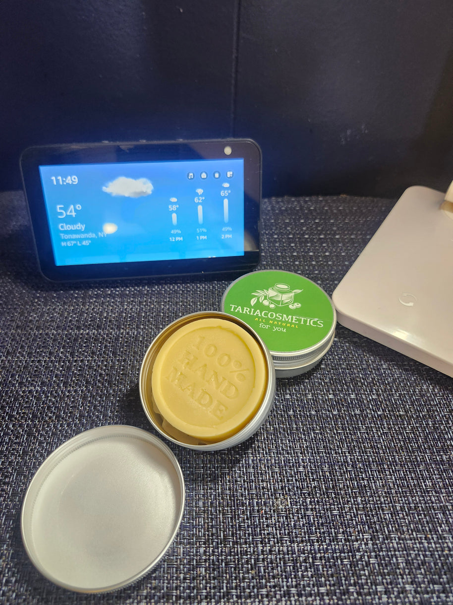 An open aluminum tin with a tan lotion bar, shown alongside it's lid and a green labeled tin near a digital display.