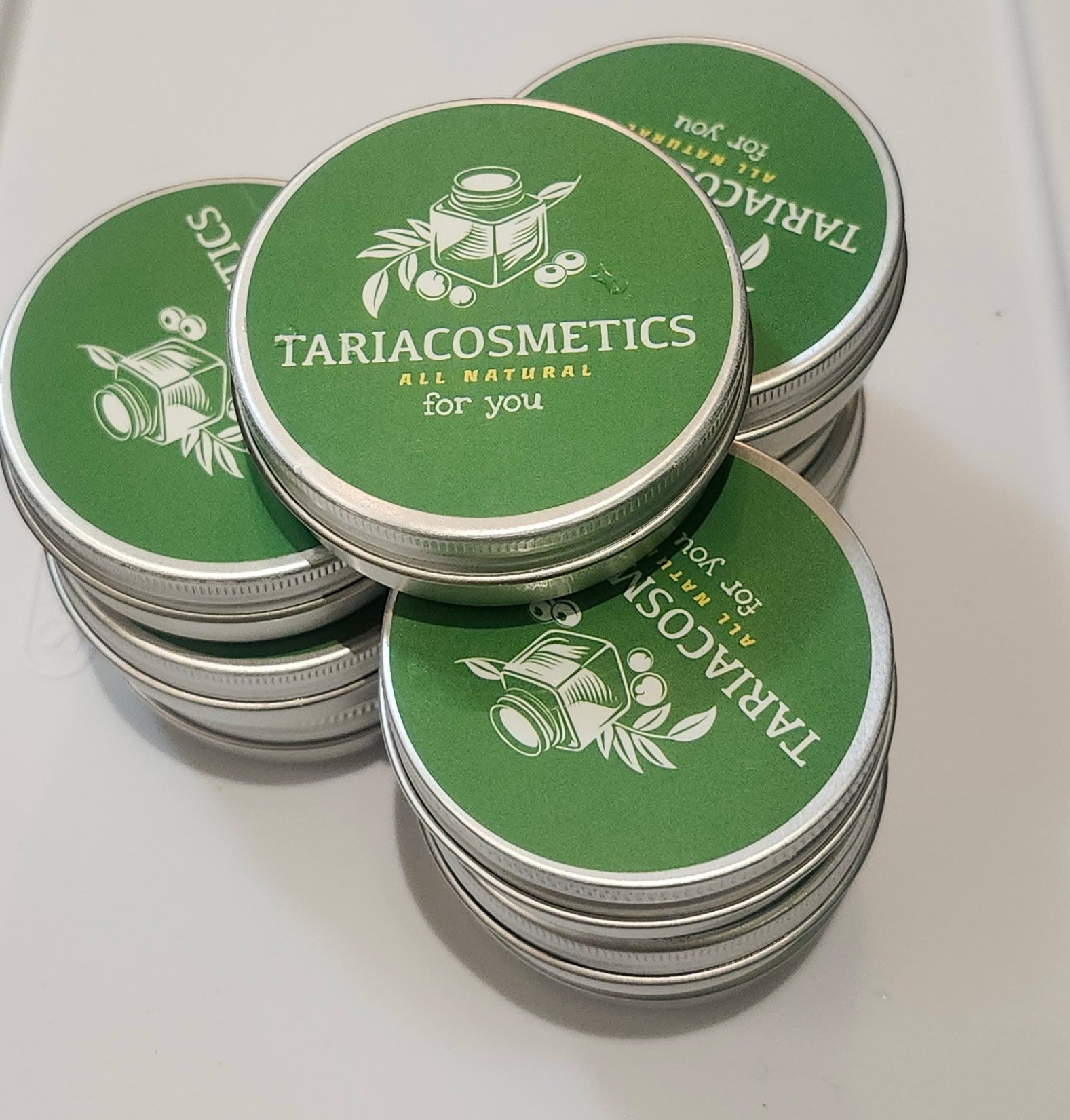 A stack of round aluminum tins featuring a green label with the branding of TariaCosmetics on them.
