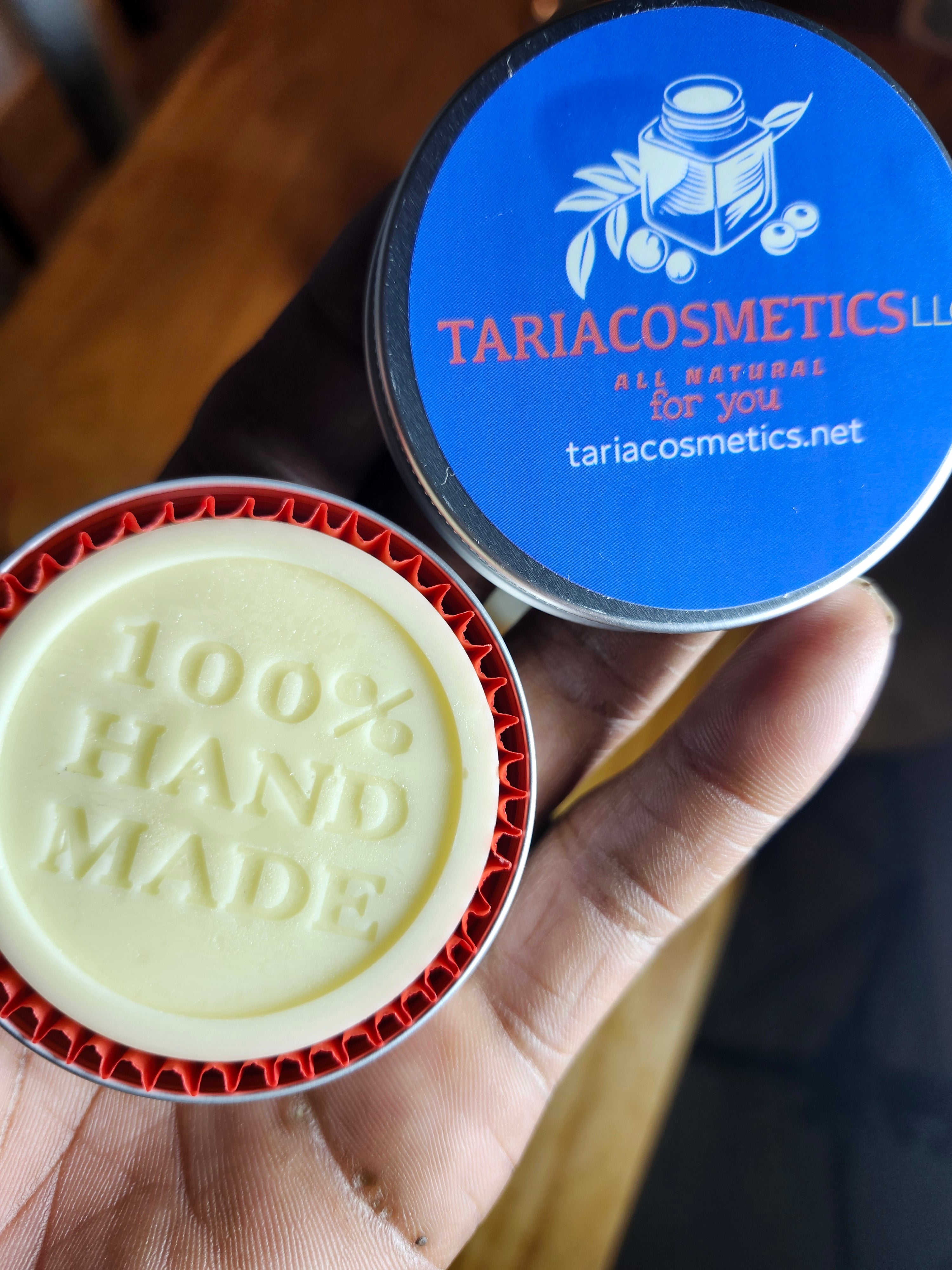 Round lotion bar with '100% handmade' branding held by a hand, next to a blue container labeled 'Taria Cosmetics'