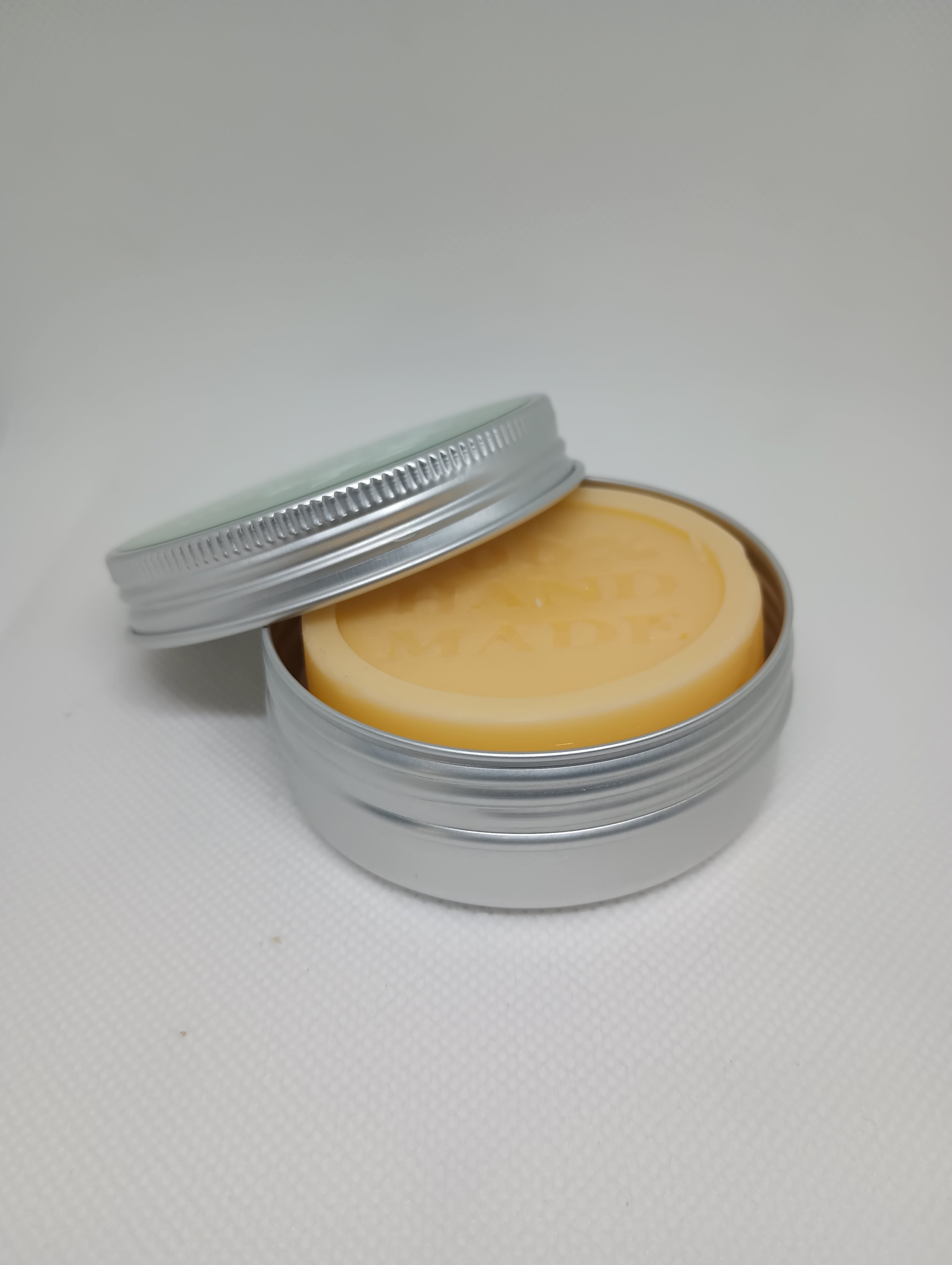 An open silver tin reveals a smooth, tan lotion bar, with the screw top lid resting partially against the side.