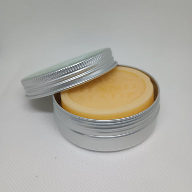 An open silver tin reveals a smooth, tan lotion bar, with the screw top lid resting partially against the side.